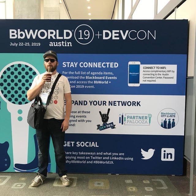 Colin Panetta drinking iced coffee standing in front of a banner with information about BBWorld.