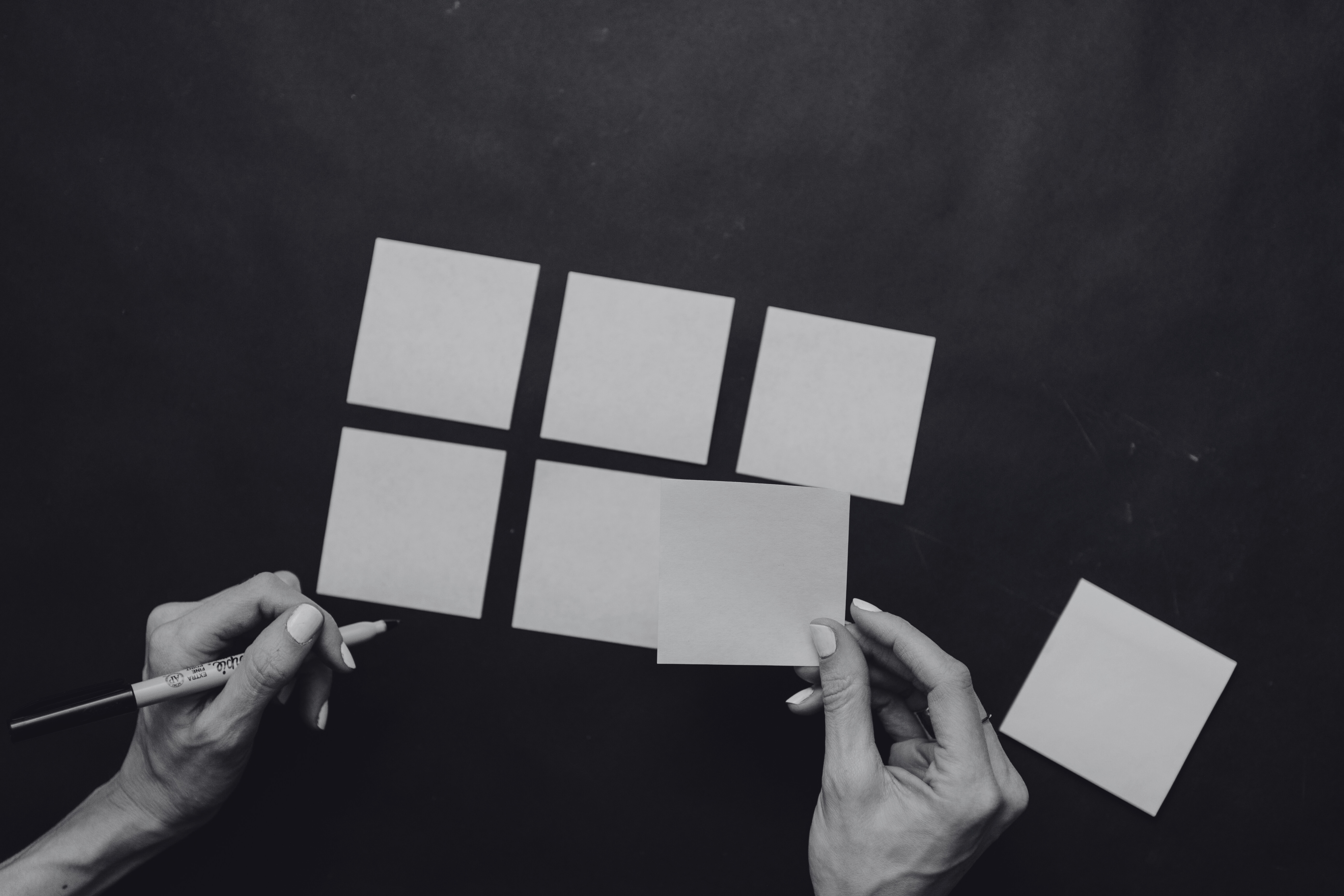 Hands arranging blank sticky notes into a grid on a dark surface, shown in black and white