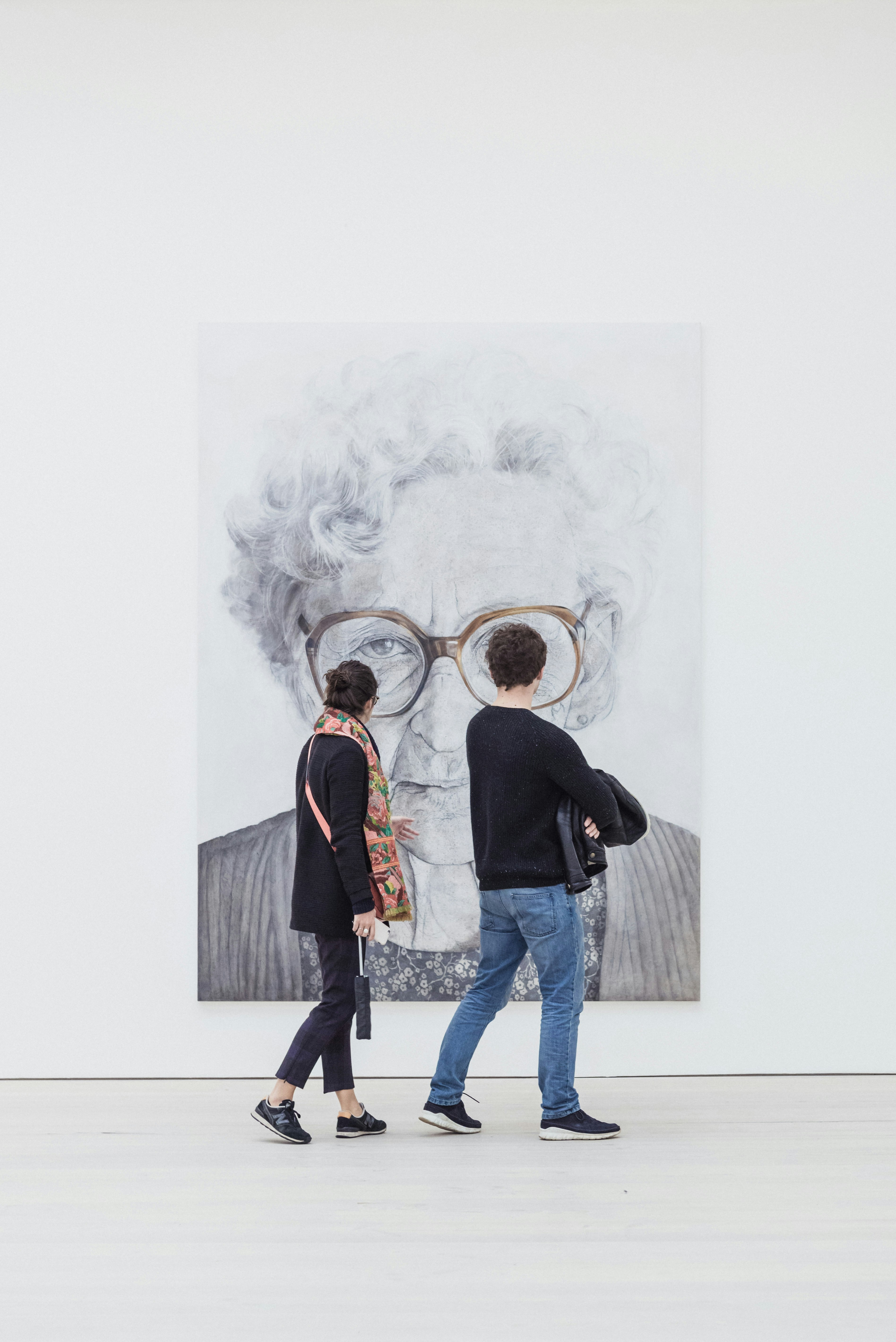 A man and woman walk through a modern art museum, viewing a large black and white portrait of an elderly woman with glasses.