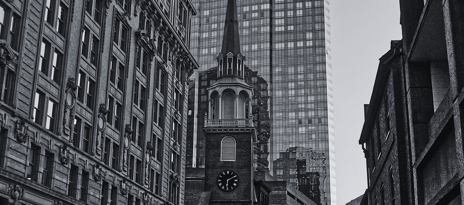 Black and white historic brick church steeple with clock tower framed by modern glass skyscraper and historic buildings in downtown Boston