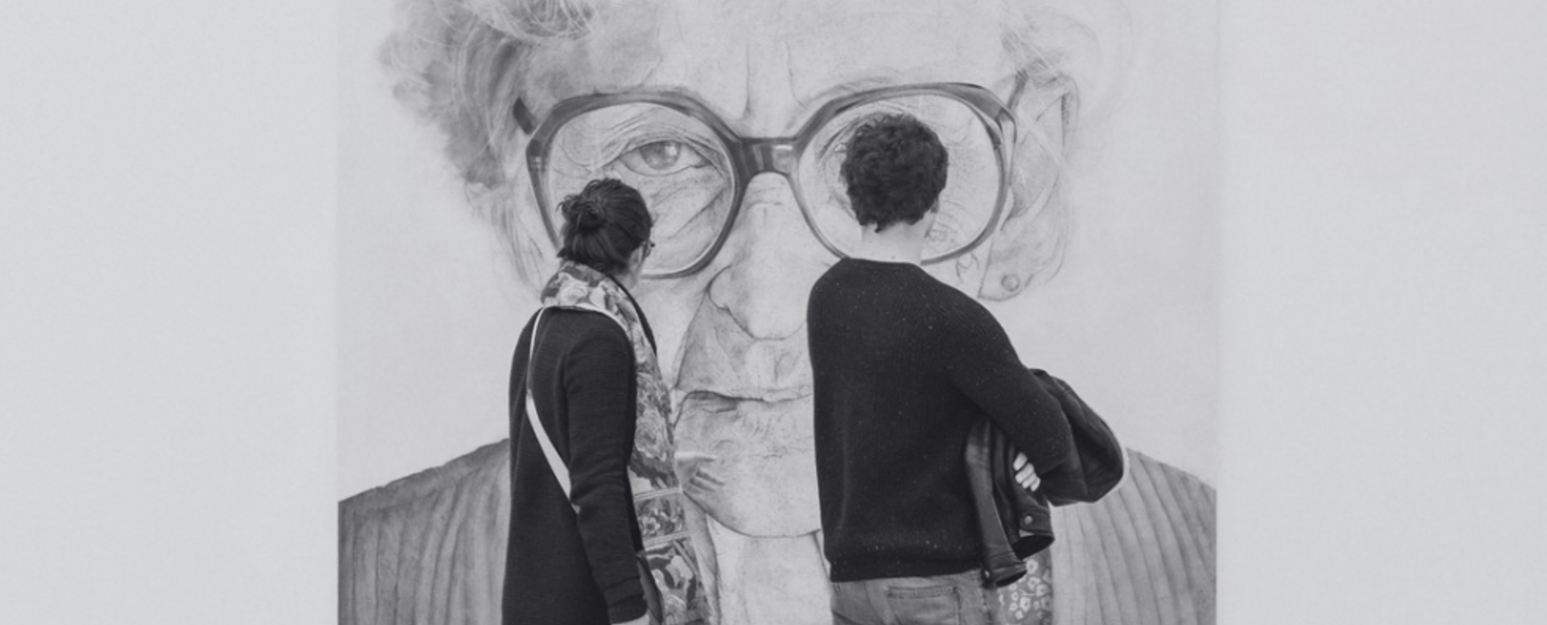 A man and woman walk through a modern art museum, viewing a large black and white portrait of an elderly woman with glasses.
