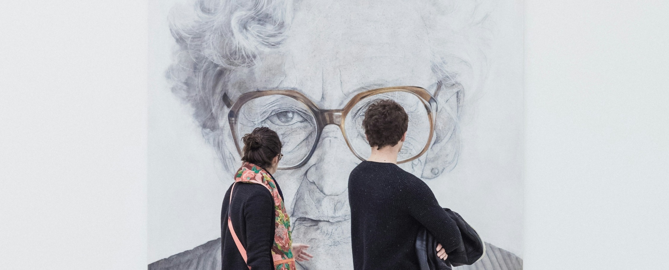 A man and woman walk through a modern art museum, viewing a large black and white portrait of an elderly woman with glasses.