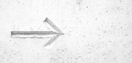 A white, textured concrete wall with a carved arrow pointing left, showing subtle shadows and cracks.