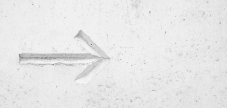 A white, textured concrete wall with a carved arrow pointing left, showing subtle shadows and cracks.
