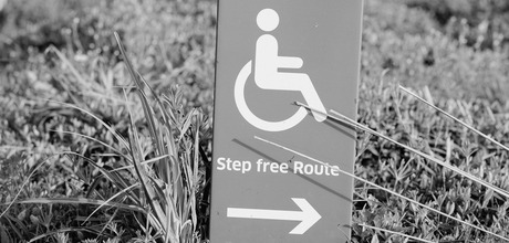A black and white image of a sign positioned in a field of grass. The sign features the International Symbol of Access and the text Step free Route