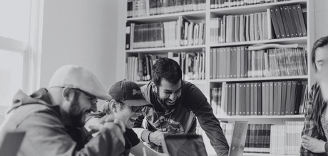 A black and white photo of a group of people huddled around a laptop at a library.