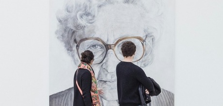A man and woman walk through a modern art museum, viewing a large black and white portrait of an elderly woman with glasses.