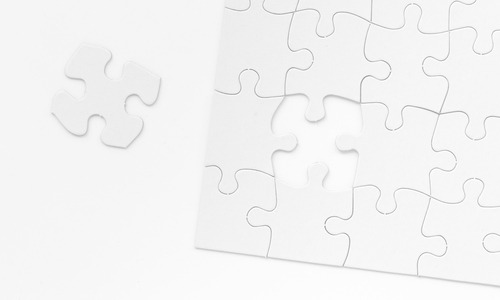 A black and white image of a puzzle missing a singular piece. The remaining piece is seen to the left of the puzzle