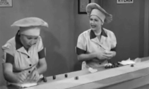 Black and white still of I Love Lucy, showing Lucille Ball and another person wearing chef outfits and standing in front of a conveyor belt