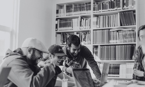 A black and white photo of a group of people huddled around a laptop at a library.