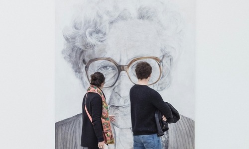 A man and woman walk through a modern art museum, viewing a large black and white portrait of an elderly woman with glasses.