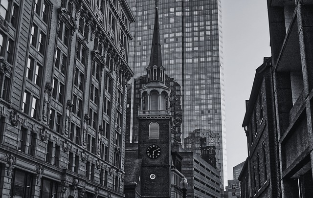 Black and white historic brick church steeple with clock tower framed by modern glass skyscraper and historic buildings in downtown Boston