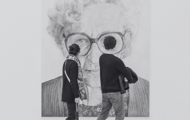 A man and woman walk through a modern art museum, viewing a large black and white portrait of an elderly woman with glasses.