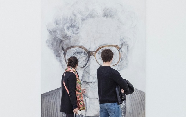 A man and woman walk through a modern art museum, viewing a large black and white portrait of an elderly woman with glasses.