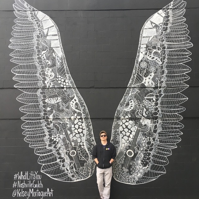 Kelly standing in front of a wall with painted wings 