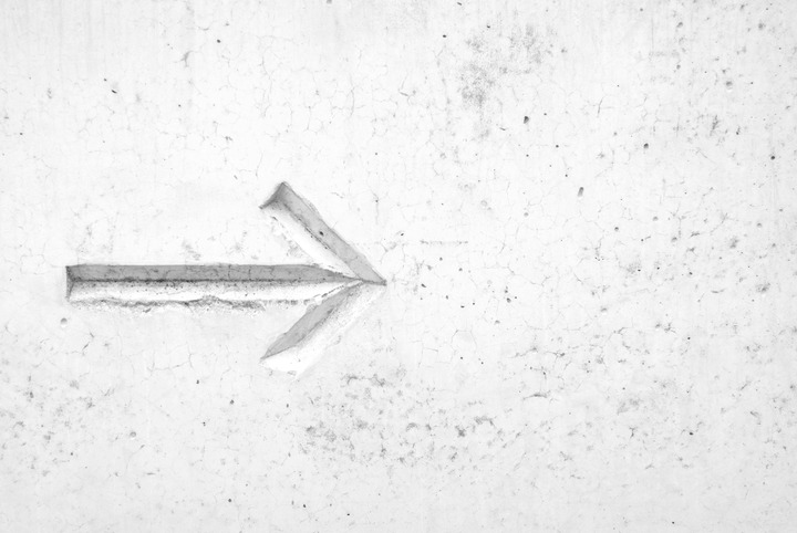 A white, textured concrete wall with a carved arrow pointing left, showing subtle shadows and cracks.