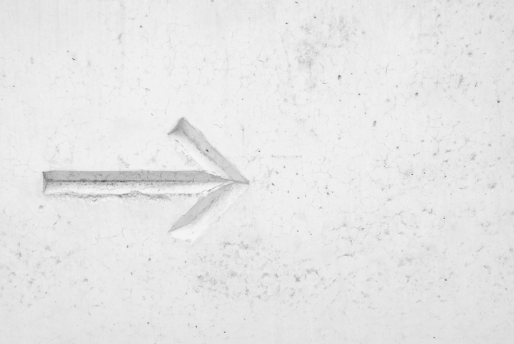 A white, textured concrete wall with a carved arrow pointing left, showing subtle shadows and cracks.