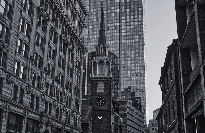 Black and white historic brick church steeple with clock tower framed by modern glass skyscraper and historic buildings in downtown Boston